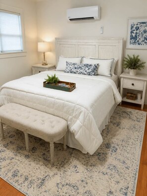 A bedroom with a large bed, a bedside table with a lamp, a small potted plant, and a wall-mounted air conditioner. - Cottage 2 - Porch Bungalow (Fairhope)