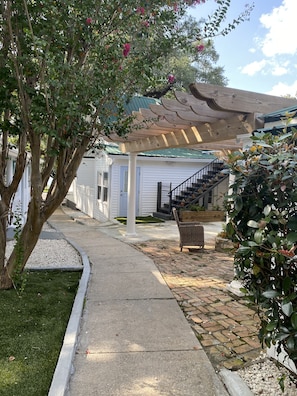 Uma área externa coberta com pérgola de madeira, uma escadaria que leva a um edifício branco e um banco.