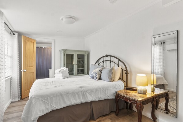 Quarto com cama, mesa de cabeceira de madeira, espelho e janela com cortinas.