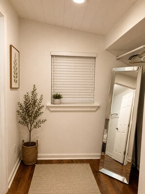 Un pasillo con piso de madera, una puerta blanca, una ventana con persianas, una planta en maceta, un cuadro enmarcado y un espejo.