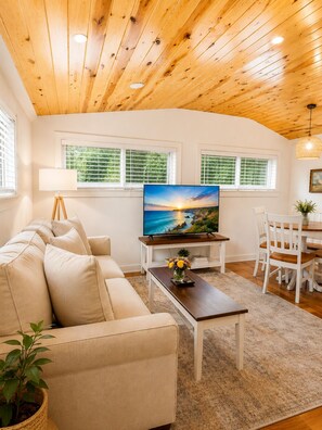 A living room with a wooden ceiling, a television on a stand, a sofa, a coffee table with flowers, and a dining area with chairs.