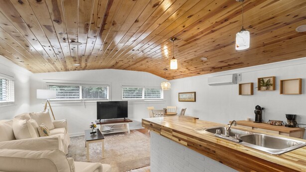 Una sala moderna con techo de madera, un televisor grande, un sofá y una cocina con cubierta de madera y lavamanos.