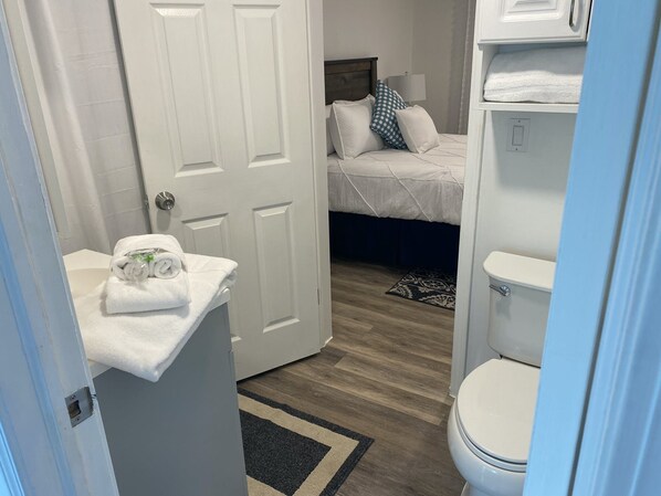 A bathroom with a white toilet, a sink with a towel, and a bed with a blue and white checkered pillow. - Pelicans Perch at The Pelicans House (Fairhope)