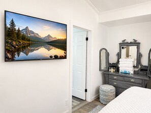 A bedroom with a large flat-screen TV mounted on the wall displaying a mountain landscape, a dark wooden dresser with a mirror, and a white door. - Pelicans Perch at The Pelicans House (Fairhope)