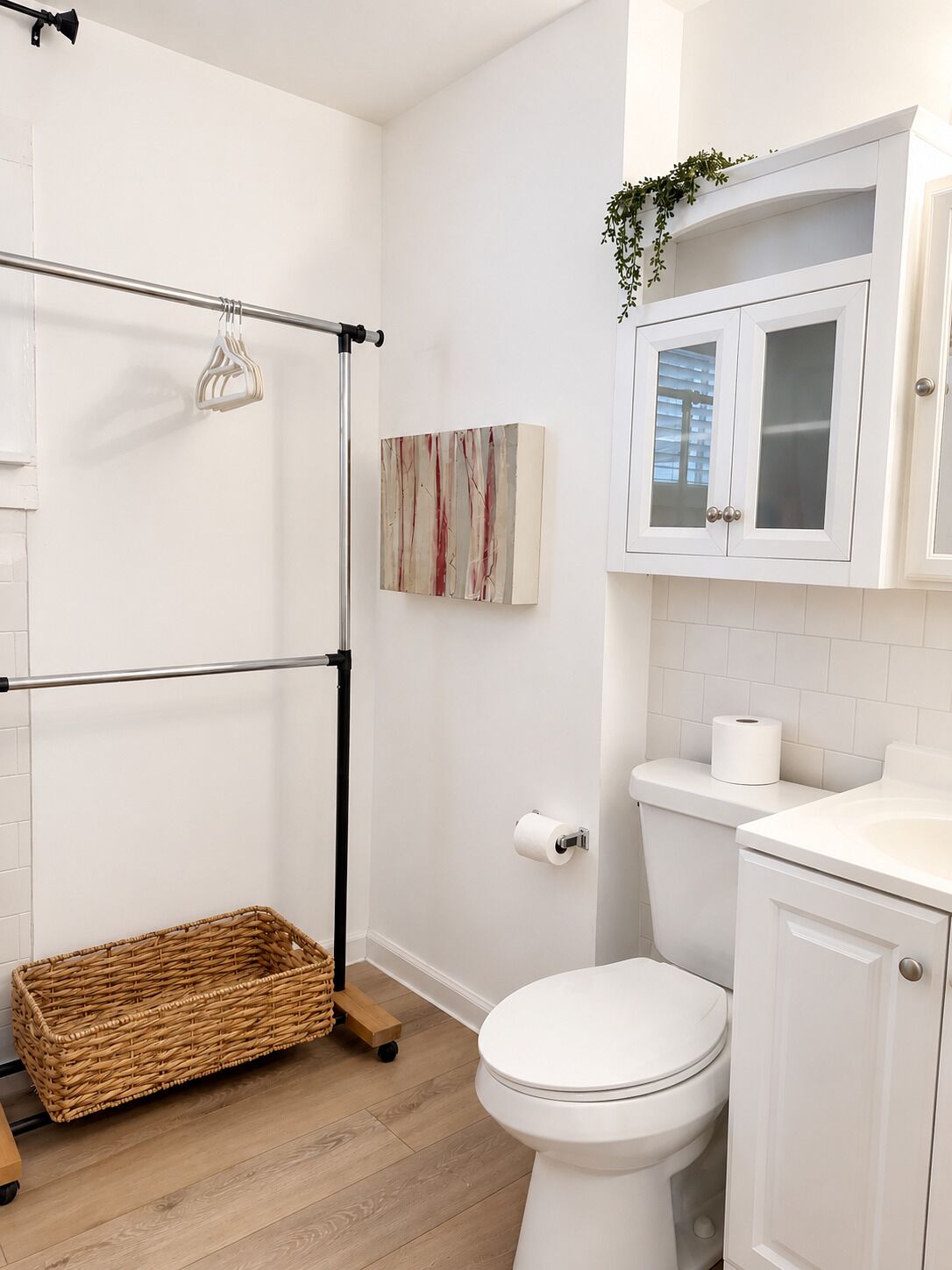 Un bagno con il water bianco, un lavabo bianco, un armadietto bianco, un appendi-asciugamani, un cesto di vimini e un quadro appeso al muro.