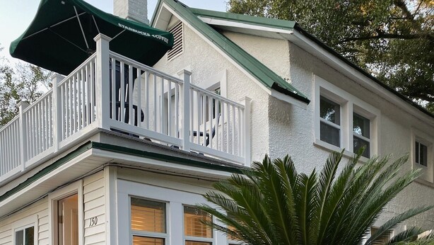 Une maison blanche à deux étages, dotée d’un balcon, avec le numéro « 150 » sur la porte et un grand palmier dans le jardin.