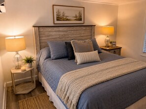 Quarto com cama, duas mesinhas de cabeceira, um criado-mudo, luminária, quadro na parede e janela com persianas.