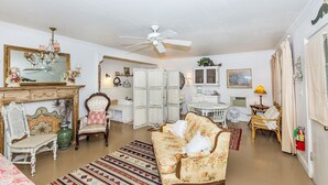 A spacious living room with a large floral sofa, a vintage armchair, and a dining table with chairs. A ceiling fan is visible, and there is a fireplace with decorative items. - Couples Getaway - 1 block to downtown- Jacuzzi tub! (Fredericksburg)