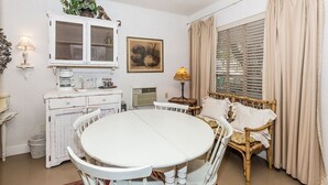 A dining area with a white round table, white chairs, a wicker sofa, a cabinet with glass doors, a painting, and a window with curtains. - Couples Getaway - 1 block to downtown- Jacuzzi tub! (Fredericksburg)