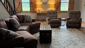 Uma sala de estar com um sofá seccional marrom, duas poltronas, uma mesa de centro e um tapete. Há um ventilador de teto, um abajur fixado na parede e uma janela com persianas de madeira.