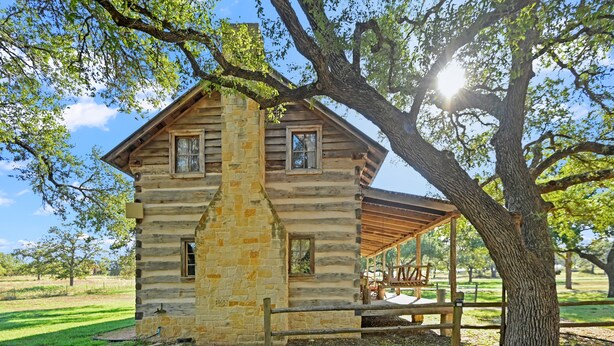 Una cabaña de troncos con chimenea de piedra, un porche y un gran árbol en primer plano.