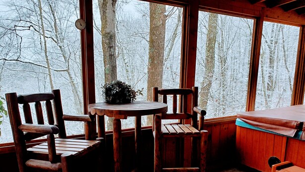 A wooden cabin interior with a round table and two chairs, a bench, and a window with a view of snow-covered trees.