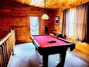 Une table de billard dans une pièce en bois, avec une porte, des fenêtres et un luminaire suspendu.