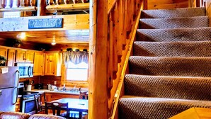 A cabin interior with a wooden staircase, a kitchen area, and a dining space.