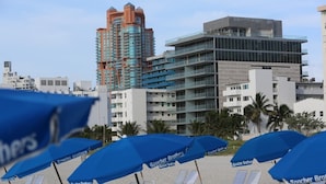 A beachside view with rows of sun loungers under blue umbrellas, a tall hotel building, and palm trees. - South Beach Ocean View King Bed + Free Beach Service (Miami Beach)