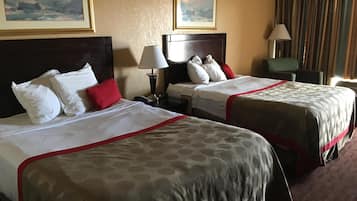 Ein Hotelzimmer mit zwei Betten, jeweils mit Kopfteil, weißer Bettwäsche und roten Akzenten. An der Wand befinden sich zwei Gemälde, neben jedem Bett eine Lampe und ein gestreifter Vorhang am Fenster.