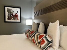 Habitación de hotel con cama, un cuadro abstracto enmarco, una lámpara de noche y un cojín decorativo estampado.