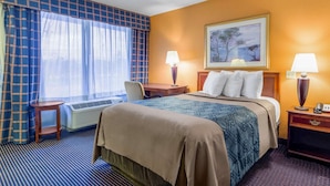 A hotel room with a bed, two bedside tables, a desk, a chair, a window with curtains, a painting on the wall, and a lamp on each bedside table. - Heritage Inn & Suites by Capital O Baton Rouge I 10 (Baton Rouge)
