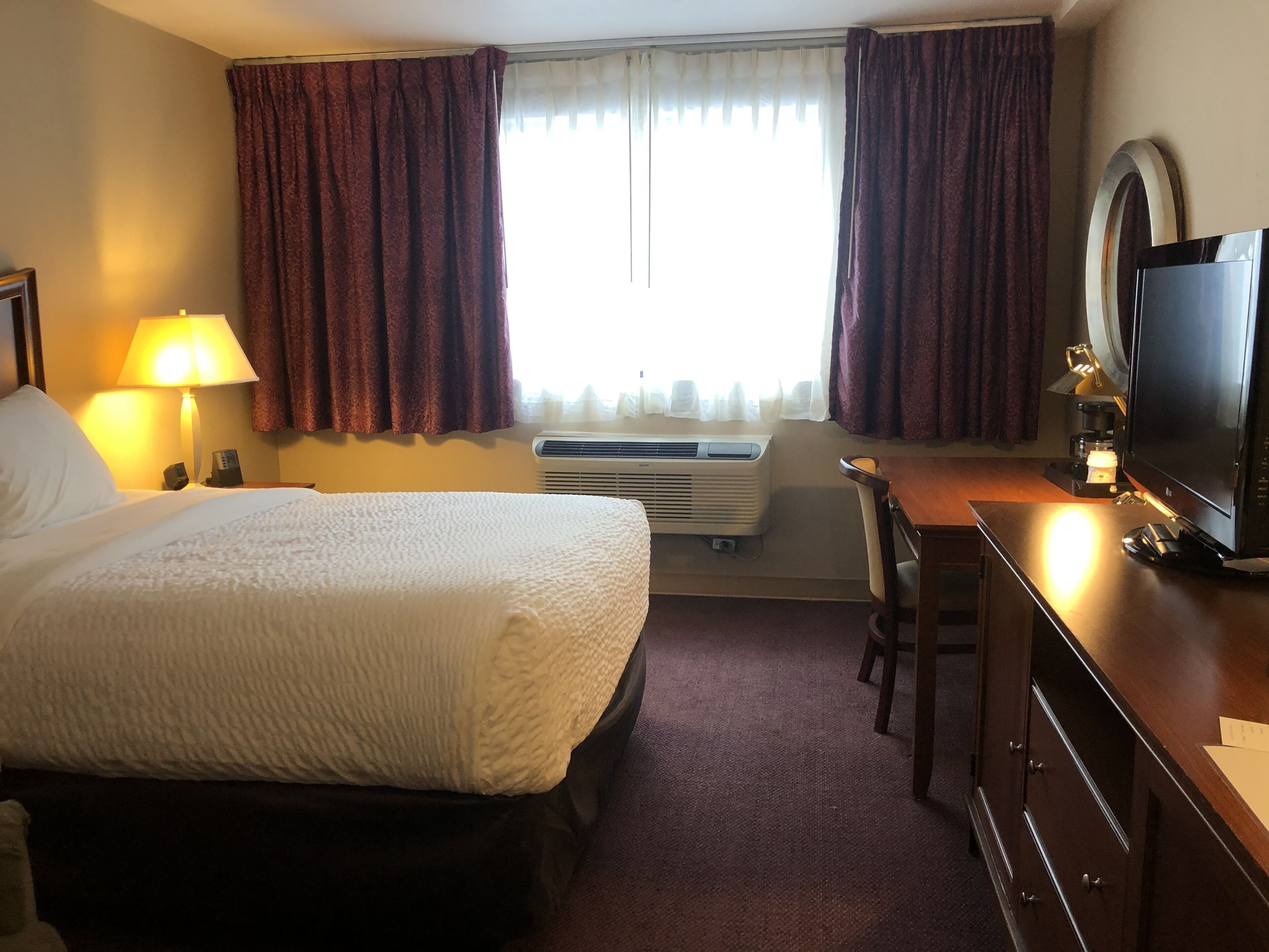 A hotel room with a bed, a desk, a television, a lamp, and an air conditioning unit.