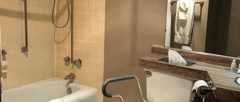 Un baño con bañera, inodoro con barra de apoyo, zona de ducha y un espejo.