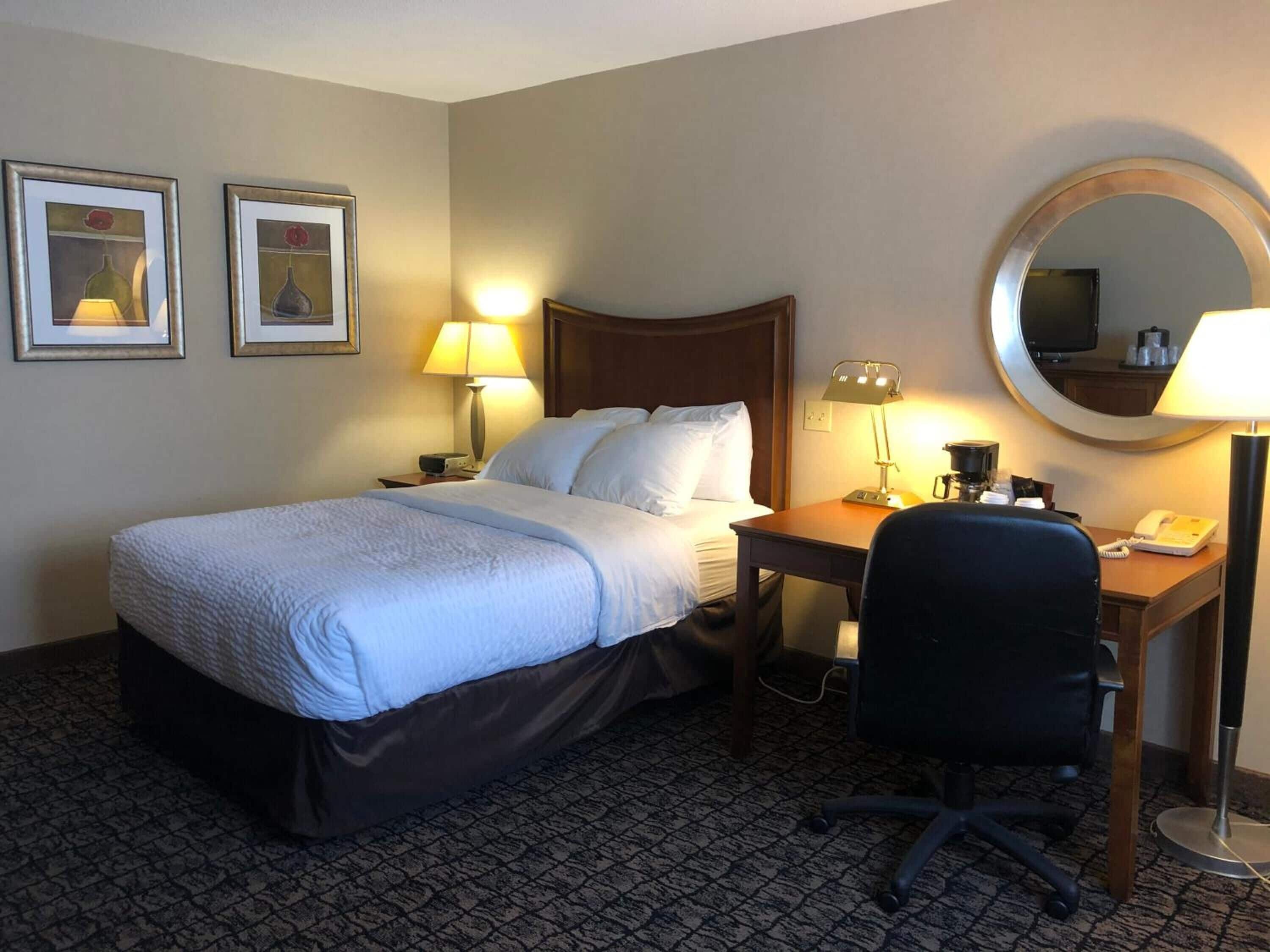 A hotel room with a bed, desk, chair, lamps, and a round mirror.