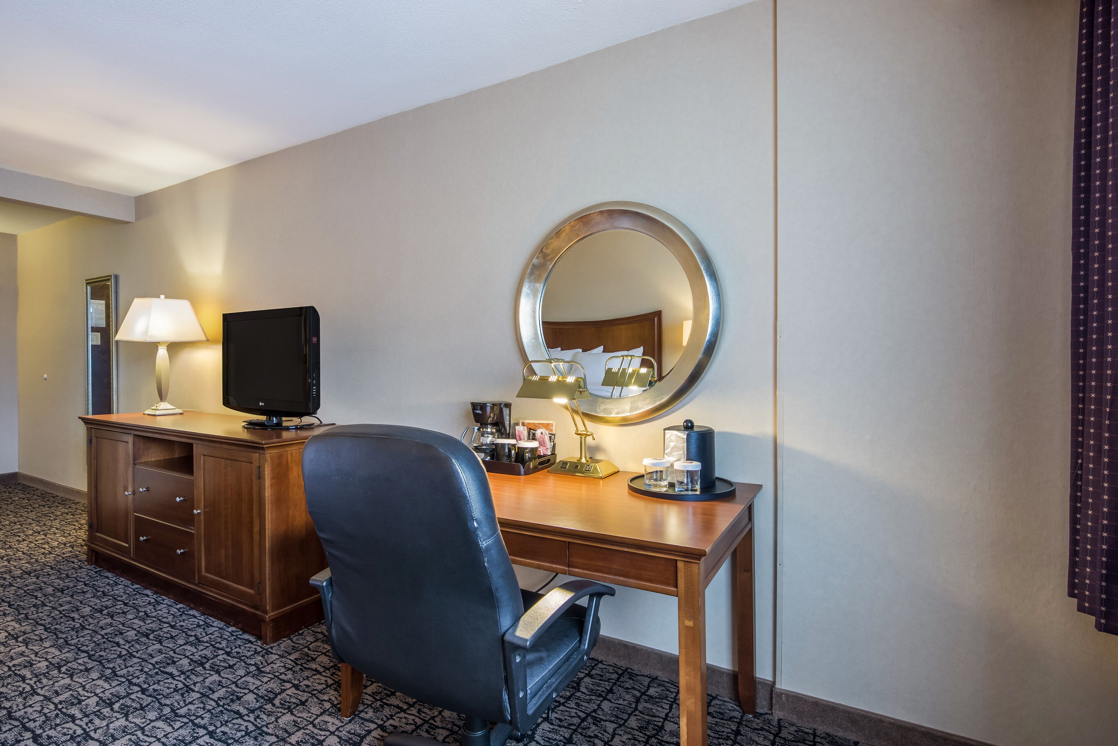 A hotel room with a wooden desk, a black office chair, a television, a lamp, a mirror, and a coffee maker.