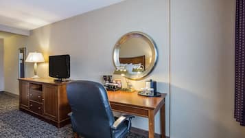 A hotel room with a wooden desk, a black office chair, a television, a lamp, a mirror, and a coffee maker.