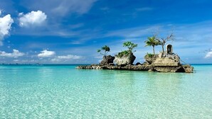 Cielo azul celeste, mar turquesa y una isla rocosa con árboles y una estructura.