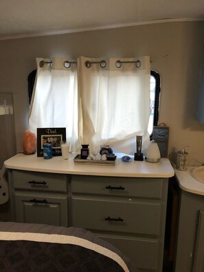Bathroom features a vanity with a single drawer and mirror, a window with a curtain, and a compact countertop with a framed photo and personal items.