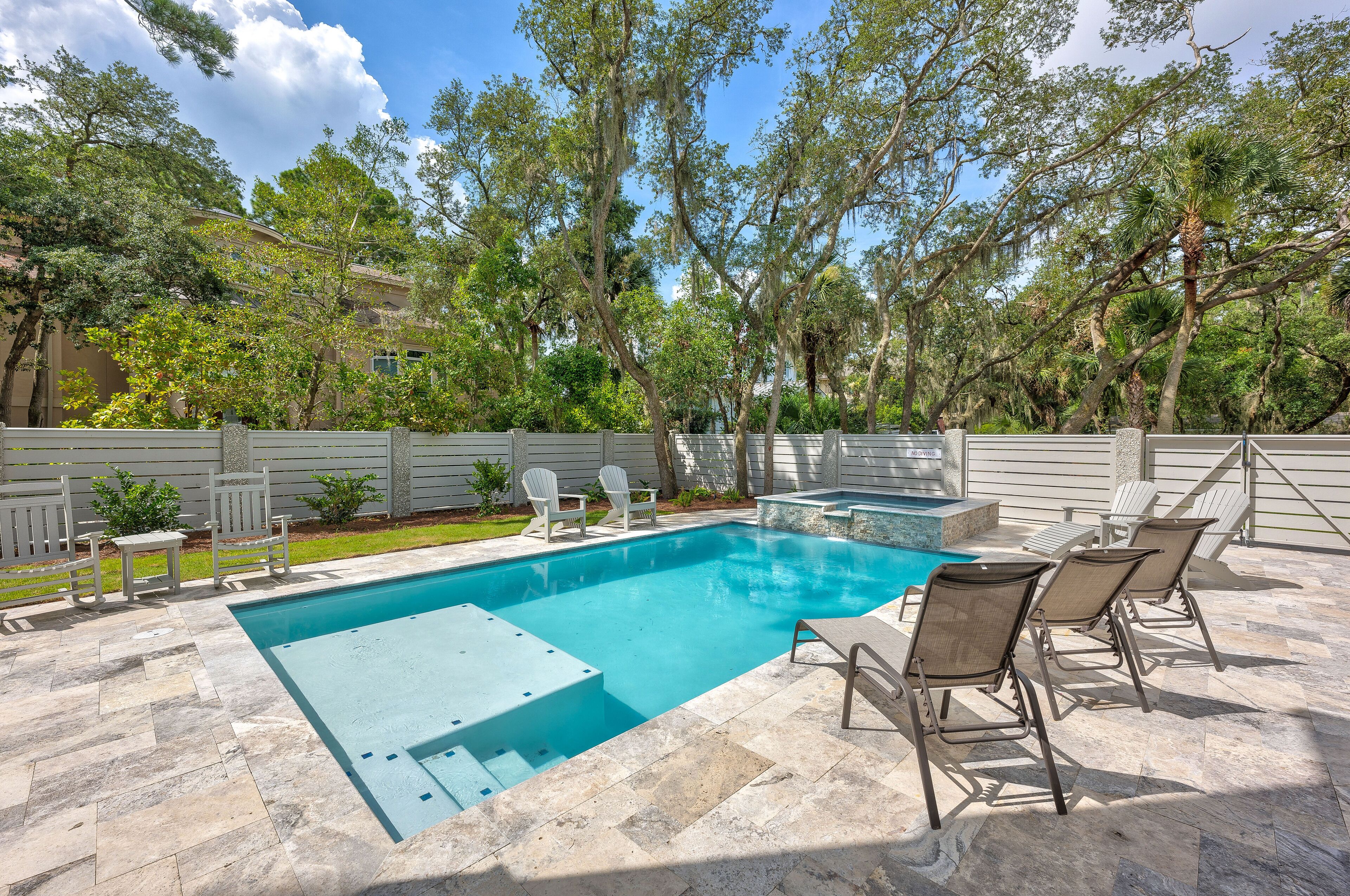 A swimming pool with a stone deck, surrounded by trees and a white fence.