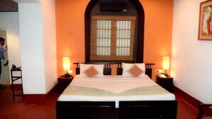 Une chambre d’hôtel avec un grand lit, deux tables de chevet, une commode, un miroir au-dessus du lit et une porte avec un cadre en bois.
