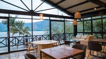 A spacious indoor area with large windows overlooking a body of water and mountains. There are multiple seating arrangements including tables and chairs.