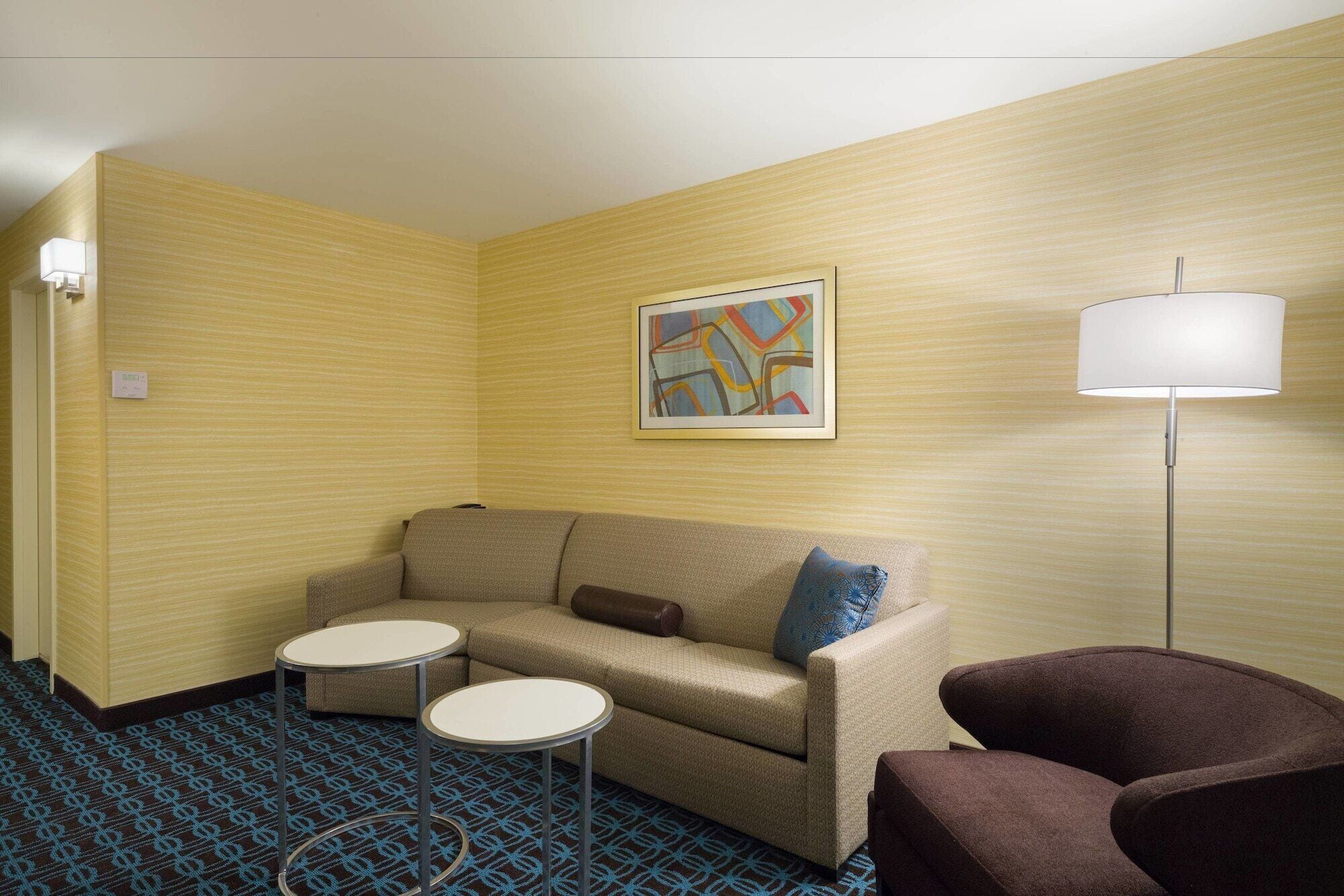 A hotel room with a beige sofa, two small round tables, a brown armchair, a floor lamp, and a wall-mounted abstract painting.
