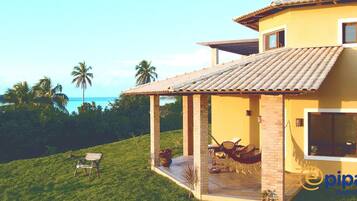 Uma casa amarela com telhado de telhas, um alpendre com mesa e cadeiras, e vista para o mar.