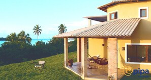 Une maison jaune avec un toit en tuiles, un patio couvert avec une table et des chaises, et une vue sur l’océan.