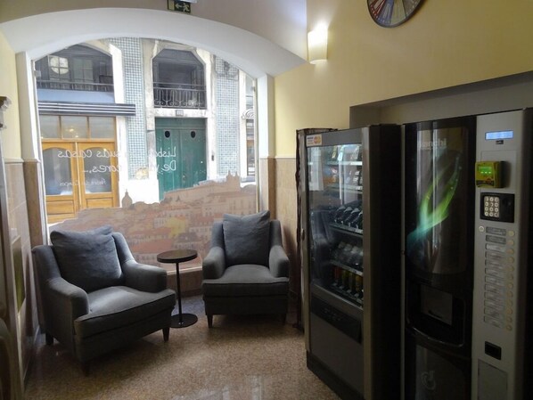 Een kamer met twee fauteuils, een bijzettafeltje, een automaat en uitzicht op een stadsgezicht door een groot raam.