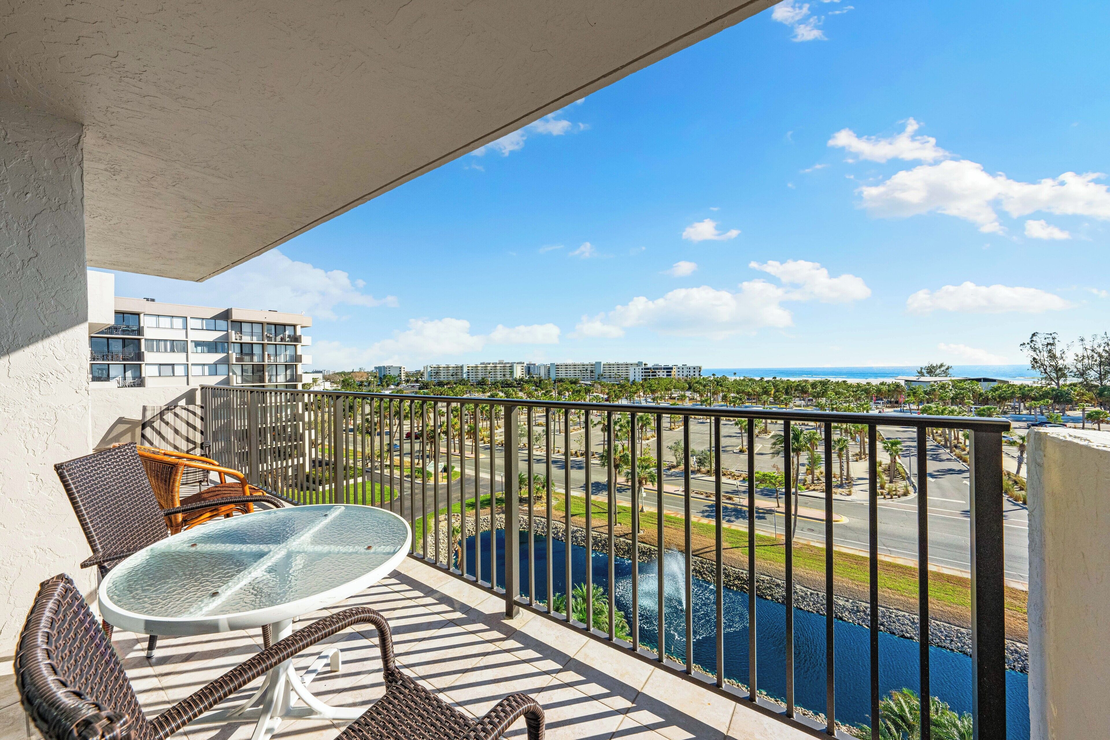 A balcony with wicker furniture, a glass table, and a view of a body of water and distant buildings.