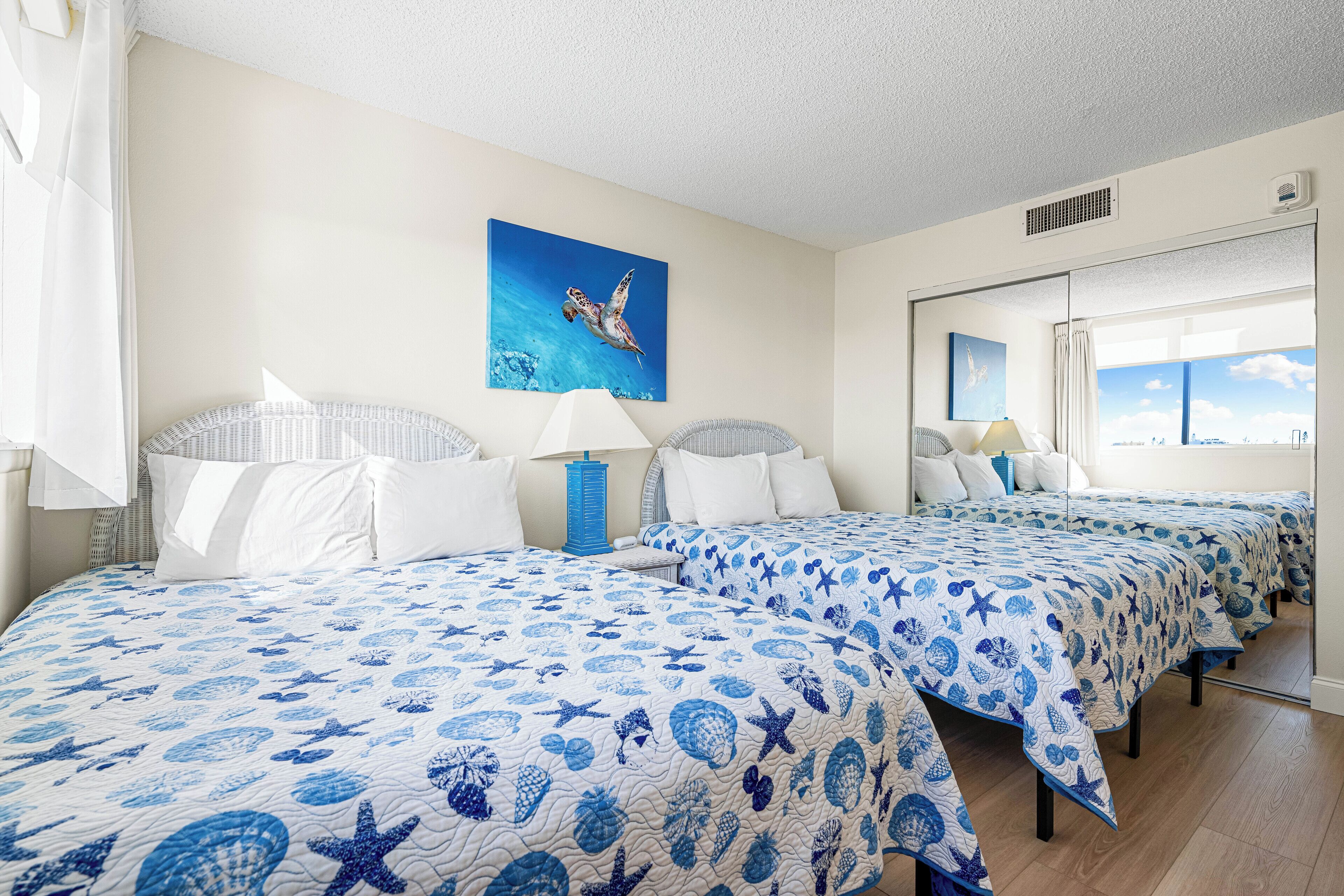 A hotel room with two beds, each with a blue and white ocean-themed bedspread, a large window, and a painting of a sea turtle on the wall.