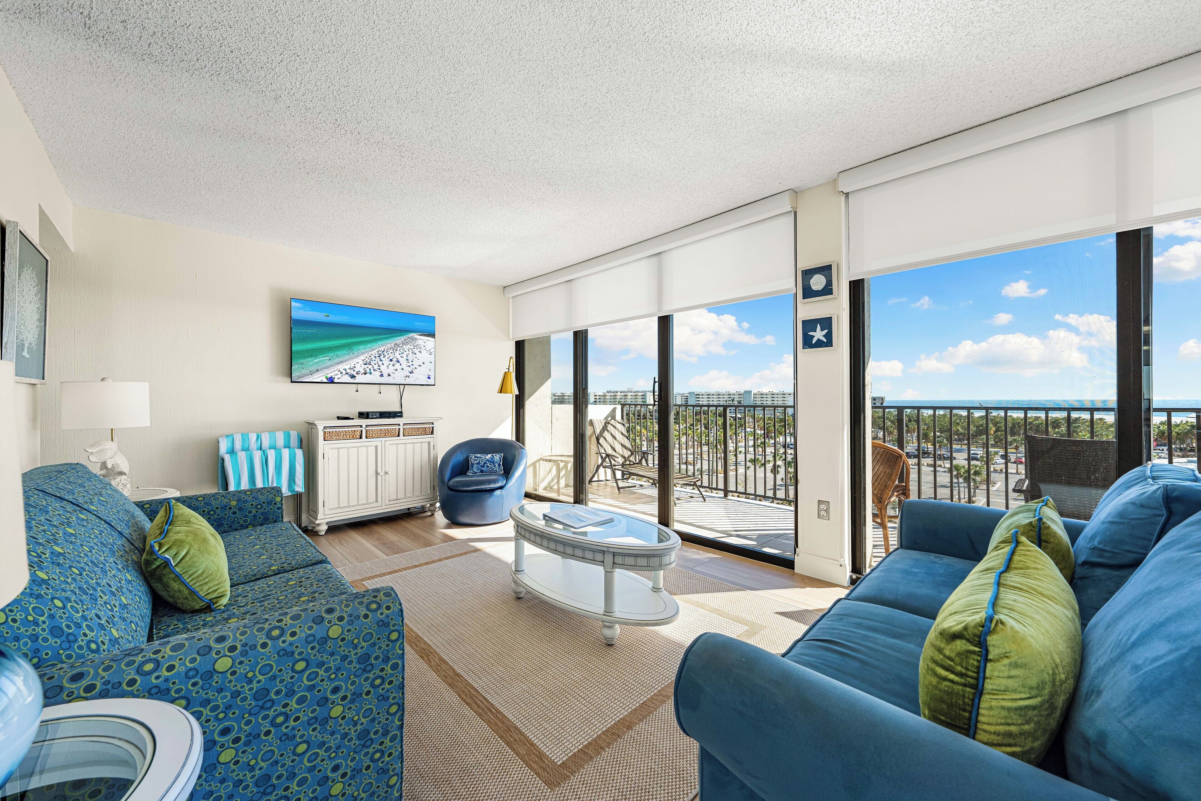 A modern living room with a large window, a flat-screen TV, and a balcony with a view of the city.