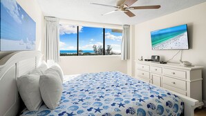 A bedroom with a large bed, a ceiling fan, a TV on the wall, and a window with a view of the beach. - Stunning 2 Bedroom/ 2 Bath with Best Siesta Key Beach Views! (Siesta Key)