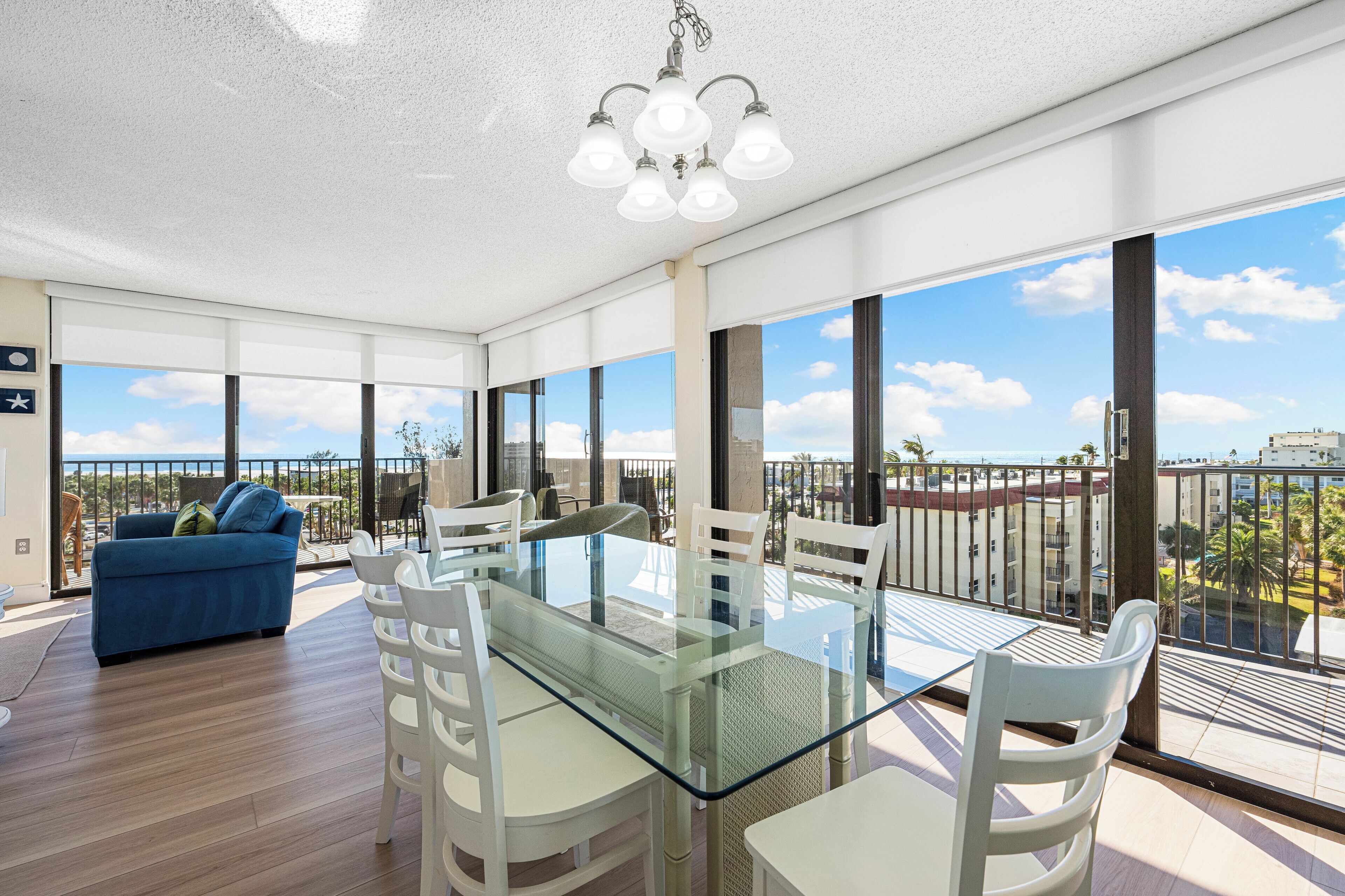 Un espace repas moderne avec une table en verre et des chaises blanches, de grandes fenêtres offrant une vue sur la ville, et un canapé bleu en arrière-plan.