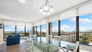 A modern dining area with a glass table and white chairs, large windows offering a view of the city, and a blue sofa in the background. - Stunning 2 Bedroom/ 2 Bath with Best Siesta Key Beach Views! (Siesta Key)
