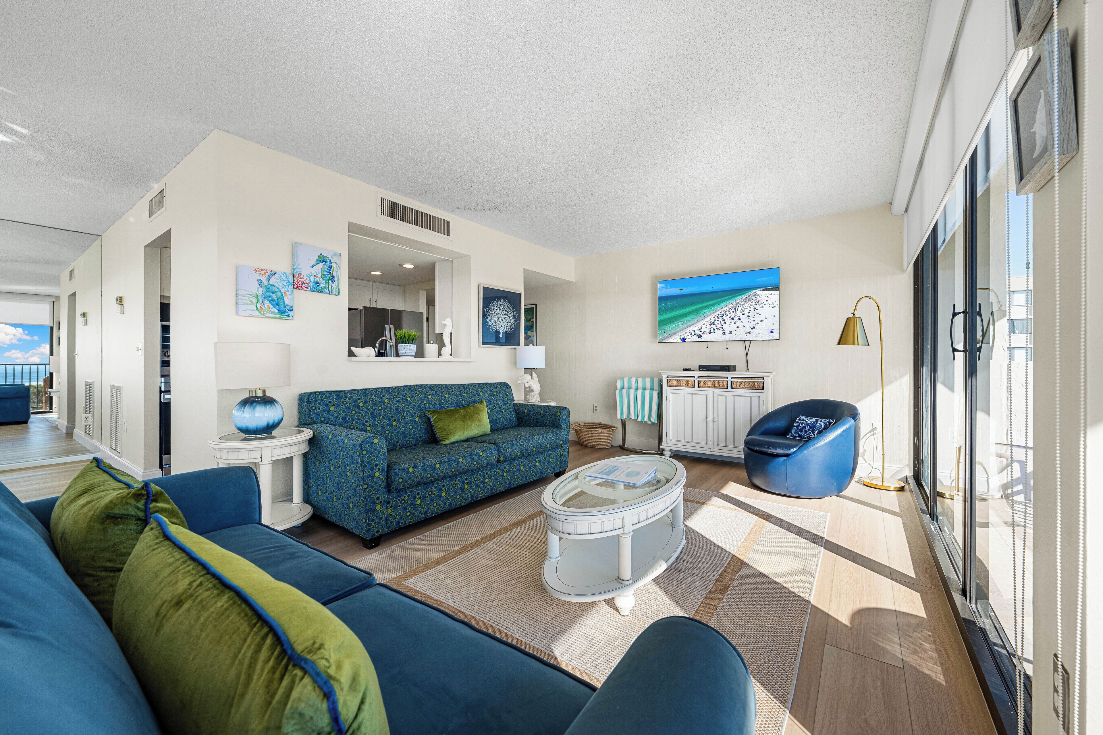 A modern living room with a blue sofa, a coffee table, and a blue armchair. There is a television mounted on the wall and a window providing natural light.