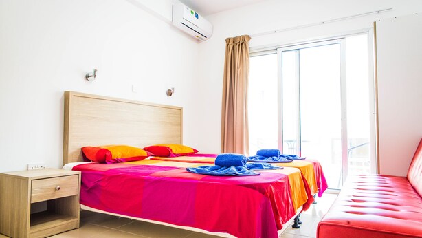 A hotel room with a bed, a nightstand, a red bench, and a sliding glass door to the balcony. - Efi Hotel Apartments (Ayia Napa)