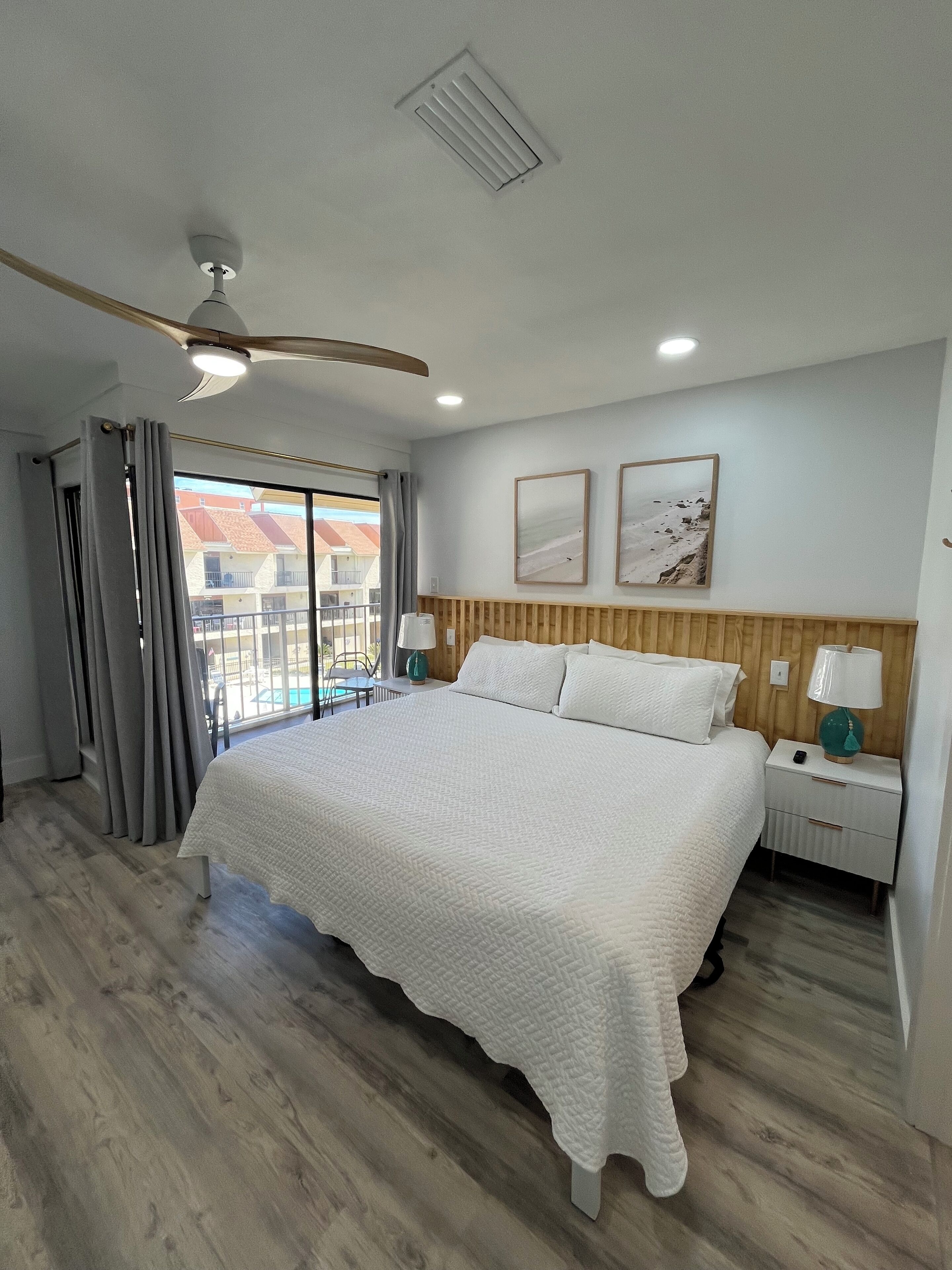 A bedroom with a large bed, wooden headboard, two bedside tables with lamps, a ceiling fan, and a view of a swimming pool outside.