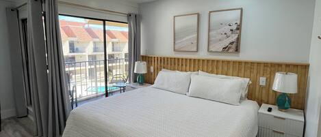 A bedroom with a large bed, wooden headboard, two bedside tables with lamps, a ceiling fan, and a view of a swimming pool outside.