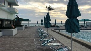 Ein Poolbereich mit Liegestühlen und Sonnenschirmen, mit Blick auf das klare Meer, und ein Gebäude mit Balkonen.
