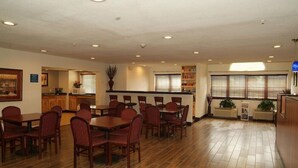 A dining area with wooden tables and chairs, a kitchen in the background, and a window with blinds.