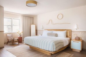 Moderni hotellihuone, jossa on puinen sänky, pieni sivupöytä, tuoli, ikkuna, jossa on verhot, ja seinään kiinnitetty valaisin.