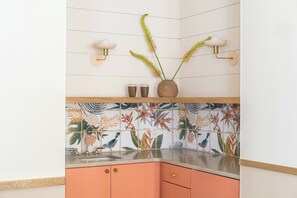 A kitchen with a tiled backsplash featuring tropical birds and leaves, a wooden countertop, and a pink cabinet with two drawers. - The Ryder Hotel (Charleston)